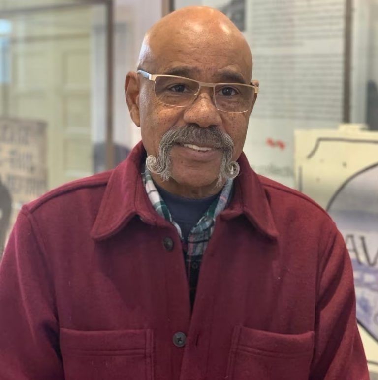 An older man in a red jacket and glasses standing in front of a wall.