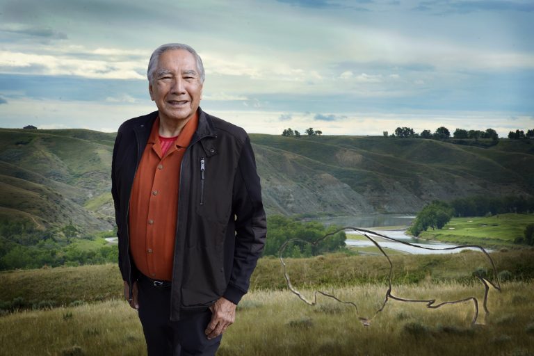 An older man smiles, standing in front of a rolling green landscape.